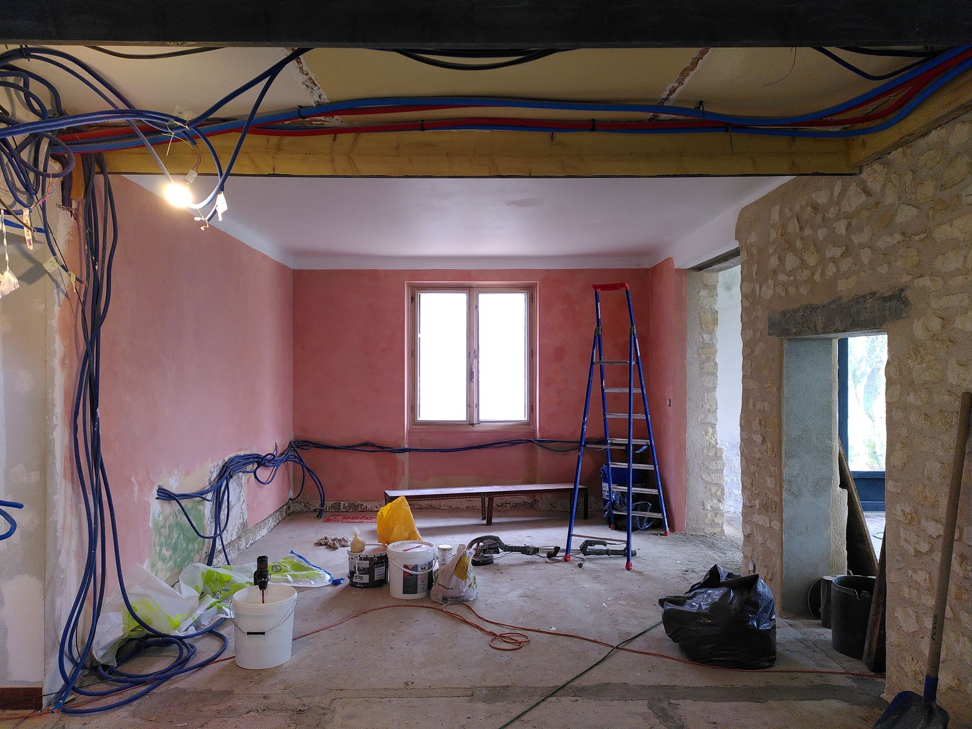 Pièce de la maison en cours de rénovation, avec murs roses, pierre apparente et gaines techniques.